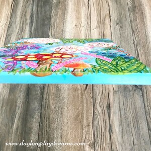 May include: A colorful painting on canvas featuring a whimsical forest scene with mushrooms, flowers, and leaves. The background is a light blue color, and the mushrooms are red with yellow spots. The painting is framed with a light blue border.