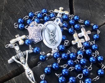 May include: A blue and silver rosary with a Saint Michael medal and a police badge charm. The rosary beads are blue and the charms are silver.