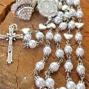 May include: A white pearl rosary with silver accents and a silver heart charm with the word "QUINCEANERA" in black lettering. The rosary also features a silver cross and a silver medal with a religious image.