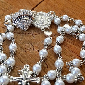 May include: White pearl rosary with a silver crucifix and a silver charm with the words "Quinceanera" in rhinestones. The charm also features a small image of the Virgin Mary.
