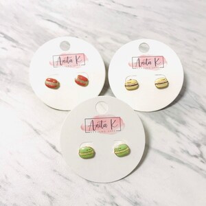 Puede incluir: Tres pares de pendientes de botón, cada uno con un diseño de macaron en miniatura. Los macarons son de color rojo, crema y verde, con una apariencia detallada. Cada par se muestra en una tarjeta blanca con el logotipo "Anita K".