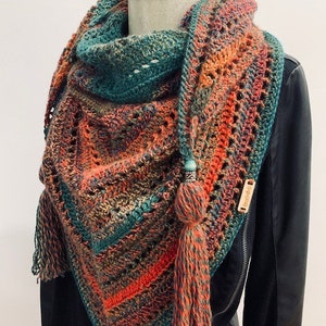 Puede incluir: Un chal de ganchillo con un patrón de rayas coloridas en tonos de verde, naranja y rojo. El chal tiene un borde de flecos y una borla en cada extremo. El chal está drapeado sobre un maniquí que lleva una chaqueta de cuero negra.