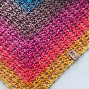 May include: A colorful crochet shawl with a rainbow ombre pattern. The shawl is made with a simple stitch and has a delicate, airy feel. The tag attached to the shawl reads "groovy and half".