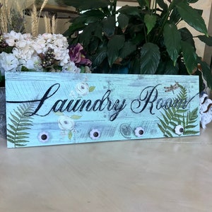 May include: A light blue wooden sign with the words "Laundry Room" in black script. The sign has a distressed look and features white flowers, green ferns, and a dragonfly. There are four white ceramic knobs with black centers attached to the sign.