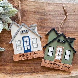 May include: Two wooden house ornaments with a white and green paint finish. The first ornament says "Kathryn & Jon Our First Home" and has the year 2024 on it. The second ornament says "The Mendenhall Family Our New Home" and has the year 2025 on it.
