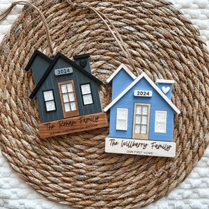 May include: Two wooden house ornaments with the year 2024 on them. The ornament on the left is painted gray and has the text "The Rohan Family" and "Our First Christmas in Our New Home". The ornament on the right is painted blue and has the text "The Willbarry Family" and "Our First Home".