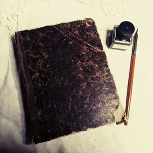 May include: A vintage, dark brown leather-bound journal with a worn and textured cover. A black inkwell and a quill pen are placed on a white surface next to the journal.