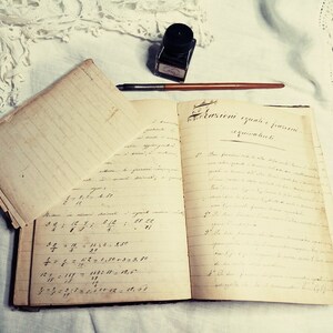 May include: An open vintage notebook with handwritten equations and text in Italian. The notebook is on a white surface with a lace doily and an inkwell with a quill pen.
