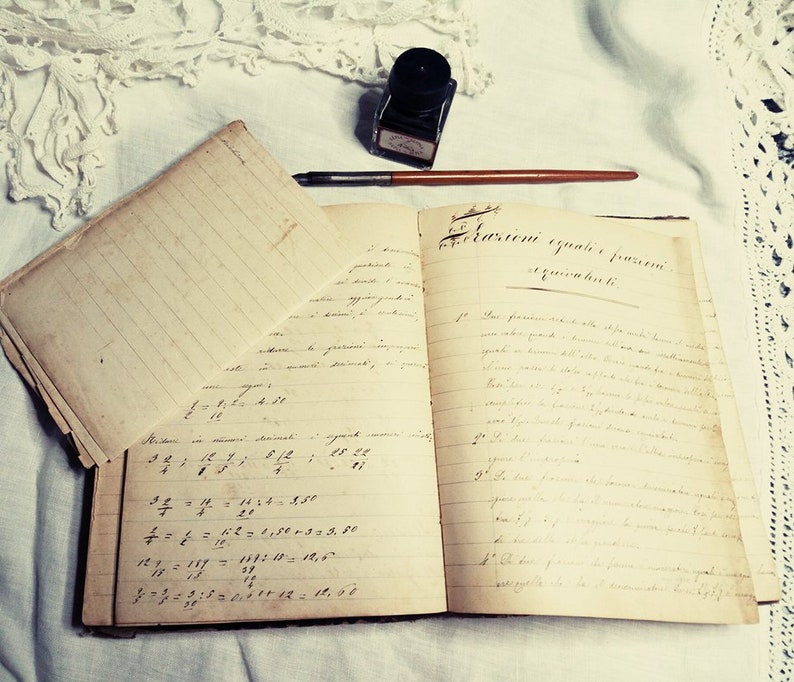 May include: An open vintage notebook with handwritten equations and text in Italian. The notebook is on a white surface with a lace doily and an inkwell with a quill pen.