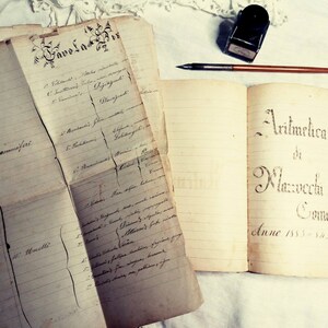 May include: An open book with the title "Aritmetica di Mazzucchelli Avvoc. Como Anno 1832." written in cursive script. The book is on a white surface with a pen and inkwell.