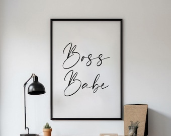 May include: A black framed print with the text "Boss Babe" in white calligraphy on a white background. The print is hanging on a white wall above a wooden desk with a black chair.