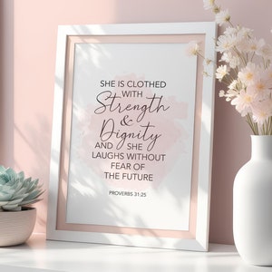 May include: A white framed print with the text "She is clothed with Strength & Dignity and she laughs without fear of the future". The frame has a pink mount. A succulent in a pot and flowers in a white vase are also present.