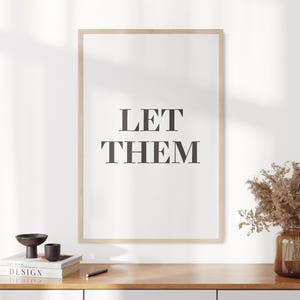 以下が含まれることがあります： 太字の濃いグレーのフォントで「LET THEM」という言葉が書かれた額入りのプリント。 プリントは明るい木製のフレームに入っており、木製の表面に表示されています。 本や花瓶など、装飾品も表面にあります。