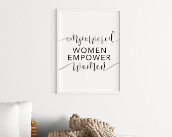 May include: A framed print with the words "empowered WOMEN EMPOWER Women" in black script. The print is in a white frame and hangs on a white wall. A beige knitted throw and white pillows are in the foreground.