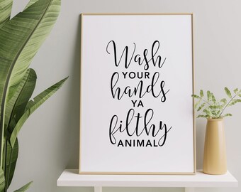 May include: A framed print with the text "Wash your hands ya filthy animal" in black script. The print is in a gold frame and sits on a white shelf. A green plant and a yellow vase with greenery are also on the shelf.