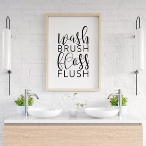 May include: A white bathroom with a framed print that says "wash brush floss flush" hanging on the wall above a double vanity with two white sinks.