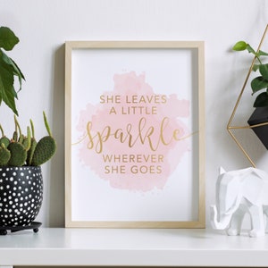 Puede incluir: Una impresión enmarcada con un fondo de acuarela rosa y letras doradas que dicen "Ella deja un poco de brillo dondequiera que vaya."