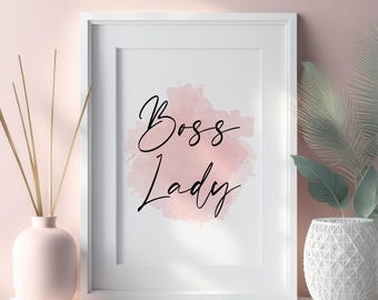 May include: A framed print with the words "Boss Lady" in elegant black script over a soft pink watercolor background. The white frame is displayed on a white surface, with a pink vase and decorative greenery in the foreground.