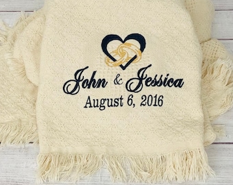 May include: Cream-colored blanket with a diamond pattern and fringe. Embroidered with a heart and two intertwined rings in navy and gold. The names "John & Jessica" and the date "August 6, 2016" are also embroidered.