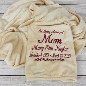 May include: A cream-colored, plush blanket with embroidered text in burgundy. The text reads "In Loving Memory of Mom Mary Etta Kaylor November 4, 1959 - March 13, 2020". The blanket is folded and draped.