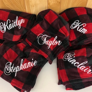 May include: Red and black buffalo plaid blankets with personalized names embroidered in white script. The names "Kaitlyn", "Taylor", "Kim", "Stephanie", and "Sinclair" are visible. The blankets are folded and stacked.