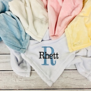 May include: A white baby blanket with the name "Rhett" embroidered in blue and black letters. The blanket is surrounded by other blankets in blue, pink, and yellow.