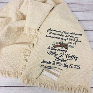 May include: Cream-colored woven blanket with fringed edges. The blanket features embroidered text and a cross design. The text reads "And the peace of God...In Loving Memory of Willis L. Coffey 'Brother' November 18, 1925 - May 27, 2019." A gift tag is attached.
