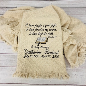 May include: A beige throw blanket with an embroidered Bible verse and a memorial message for Catherine Brutout. The verse reads "I have fought a good fight, I have finished my course, I have kept the faith. II Timothy 4:7". The memorial message reads "In Loving Memory of Catherine Brutout July 10, 1950-April 17, 2020".