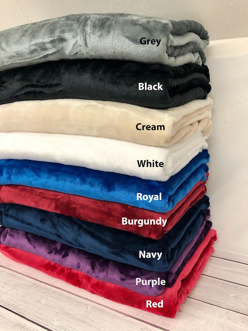 May include: A stack of ten plush blankets in various colors, including grey, black, cream, white, royal blue, burgundy, navy, purple, and red. Each blanket is folded and stacked on top of the other, with the color name written below each blanket.