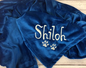 Pode incluir: Um cobertor azul royal de pelúcia com o nome "Shiloh" bordado em branco. Duas pegadas prateadas são bordadas abaixo do nome. O cobertor está dobrado, mostrando sua textura macia.