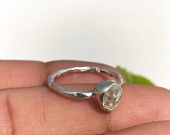 Op de afbeelding: Een zilveren ring met een facetgeslepen, heldere edelsteen. De ring heeft een getextureerde band en een bezel setting voor de edelsteen. De ring wordt op een vinger getoond. Het is een sieraad.