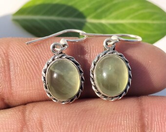 May include: A pair of silver earrings with oval, pale green gemstones. Each earring features a twisted rope design around the gemstone and a simple hook closure. The earrings are displayed against a blurred background.