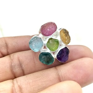 May include: A silver ring with a floral design, set with a cluster of colourful gemstones. The ring features raw, uncut stones in shades of pink, blue, green, yellow, and purple. The gemstones are set in individual silver settings, creating a flower-like arrangement.