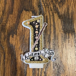 May include: A gold glitter candle shaped like the number one with a black wand and a banner that says "Lumos".