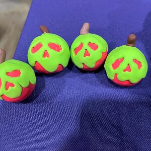 May include: Four red and green apple-shaped figurines with a skull design. The apples have a red base, a green top, and a brown stem. The figurines are arranged on a purple surface.