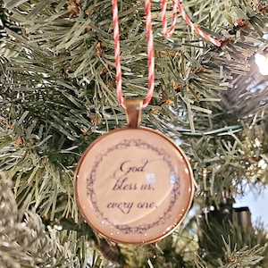 May include: A round glass ornament with a gold trim and a white background. The ornament has a cursive inscription that reads "God bless us, every one."