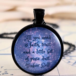 May include: A round black metal pendant necklace featuring a blue and purple galaxy-themed background with pink sparkles. The text reads: All you need is faith, trust, and a little bit of pixie dust. Tinker Bell. The pendant hangs on a black chain.