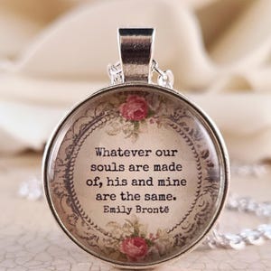 May include: A round silver pendant necklace featuring a quote by Emily Brontë that reads, Whatever our souls are made of, his and mine are the same. The text is centered on a beige background with a decorative border and small pink rose illustrations.