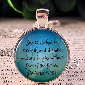 May include: A round silver pendant necklace featuring a blue and green watercolor background with black text that reads: She is clothed in strength and dignity, and she laughs without fear of the future. Proverbs 31:25. The pendant has a glass dome.