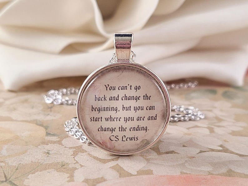 Puede incluir: Un colgante plateado redondo con una cita de C.S. Lewis. La cita dice: "No puedes volver atr&aacute;s y cambiar el principio, pero puedes empezar donde est&aacute;s y cambiar el final." Fondo floral.