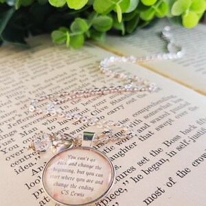 Puede incluir: Collar con colgante plateado con un dije redondo. El dije presenta una cita de C.S. Lewis sobre un fondo floral. El collar descansa sobre un libro abierto, con follaje verde en el fondo.