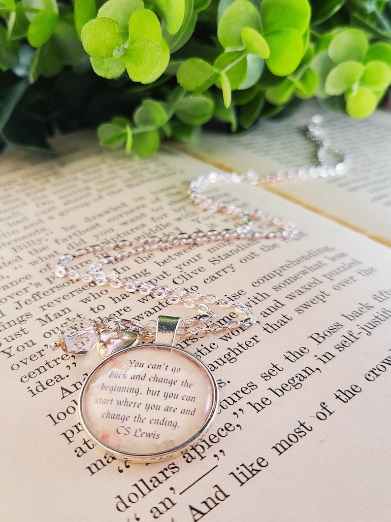 Puede incluir: Collar con colgante plateado con un dije redondo. El dije presenta una cita de C.S. Lewis sobre un fondo floral. El collar descansa sobre un libro abierto, con follaje verde en el fondo.