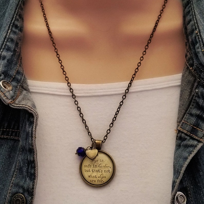 May include: A bronze necklace with a round pendant that reads "A ship is safe in harbor, but that's not what ships are for." The pendant is accented with a small heart charm and a blue glass bead.