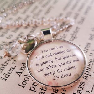 Puede incluir: Un collar con colgante plateado con un colgante redondo de cristal transparente. El colgante presenta una cita de C.S. Lewis sobre un fondo floral. El collar descansa sobre un libro abierto con texto impreso.