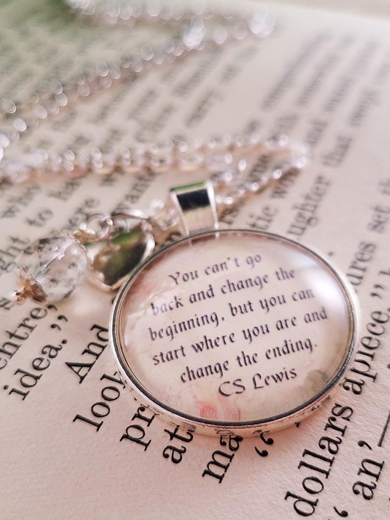 Puede incluir: Un collar con colgante plateado con un colgante redondo de cristal transparente. El colgante presenta una cita de C.S. Lewis sobre un fondo floral. El collar descansa sobre un libro abierto con texto impreso.