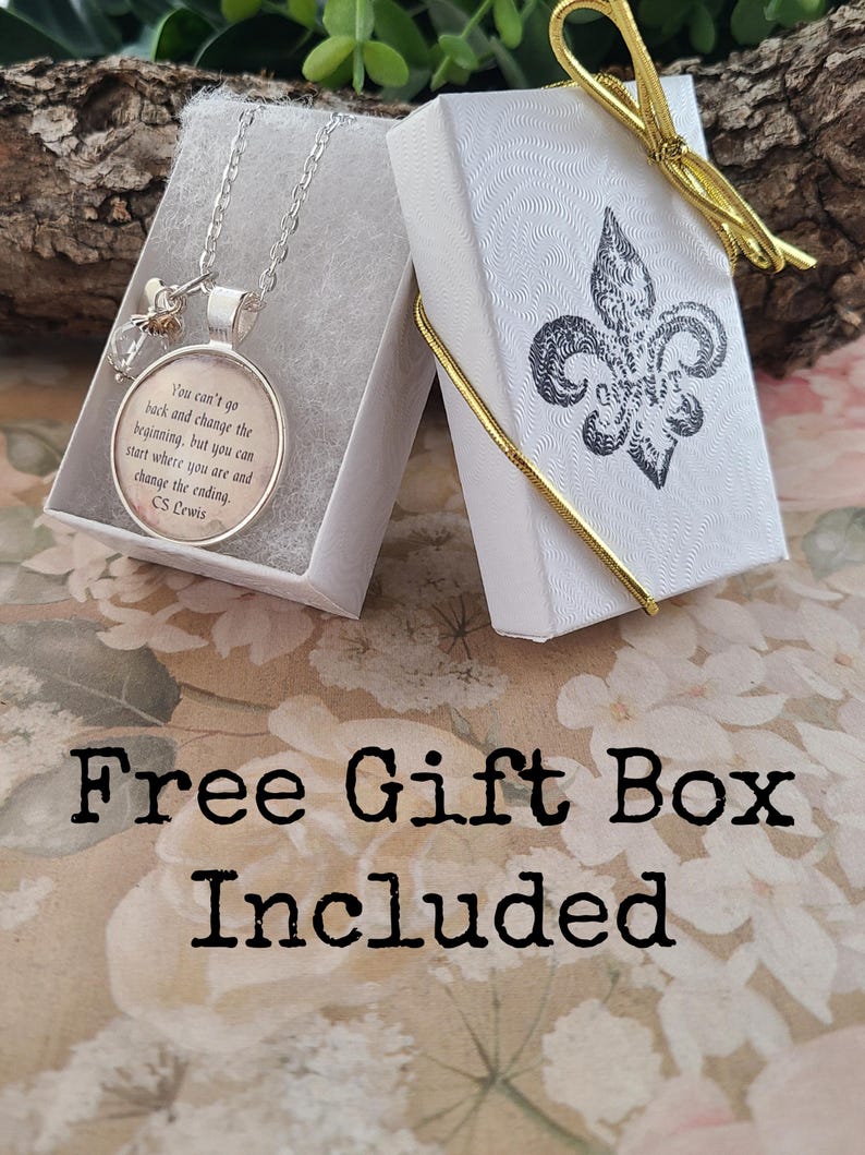 Puede incluir: Un collar con colgante plateado con una cita de C.S. Lewis dentro de un colgante redondo, presentado en una caja de regalo blanca abierta. La caja est&aacute; decorada con un dise&ntilde;o de flor de lis y una cinta dorada. El texto "Free Gift Box Included" tambi&eacute;n es visible.