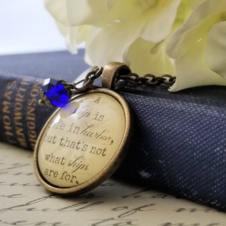 May include: A gold necklace with a round pendant featuring a white background and black text that reads "A ship is safe in harbor, but that's not what ships are for." The pendant has a blue crystal charm attached to the chain.