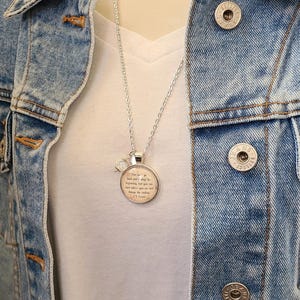 Puede incluir: Collar plateado con un colgante redondo con una cita y un peque&ntilde;o dije de cristal. El collar se lleva sobre una camiseta blanca y una chaqueta vaquera.