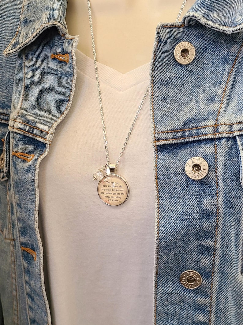 Puede incluir: Collar plateado con un colgante redondo con una cita y un peque&ntilde;o dije de cristal. El collar se lleva sobre una camiseta blanca y una chaqueta vaquera.