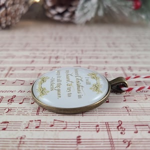 Pu&ograve; includere: Un ciondolo rotondo con una cornice in metallo color bronzo, sfondo bianco e testo dorato che recita: I will honor Christmas in my heart & try to keep it all the year. -Dickens. Il ciondolo &egrave; decorato con illustrazioni di agrifoglio dorato.